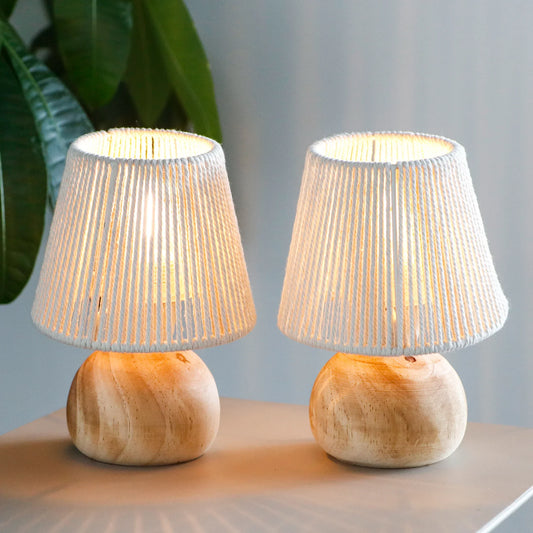 Two matching table lamps with woven off-white conical shades and rounded natural wood bases, shown lit on a tabletop with a blurred indoor background and green leaves at the left edge.